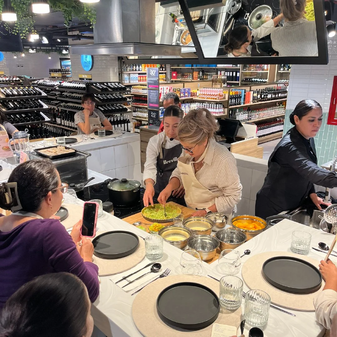 Grupo de personas participando en una clase de cocina en una tienda, preparando alimentos en una mesa con utensilios y ingredientes.