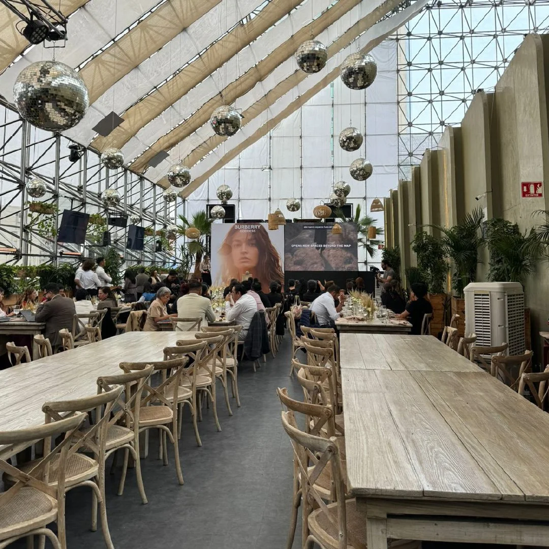 Un gran salón con techo alto de estructura metálica y paredes de cristal. Representaciones de espejos disco colgados del techo. Mesas de madera y sillas de madera con asientos de mimbre, ocupadas por varias personas. Algunas plantas decorativas. Pant