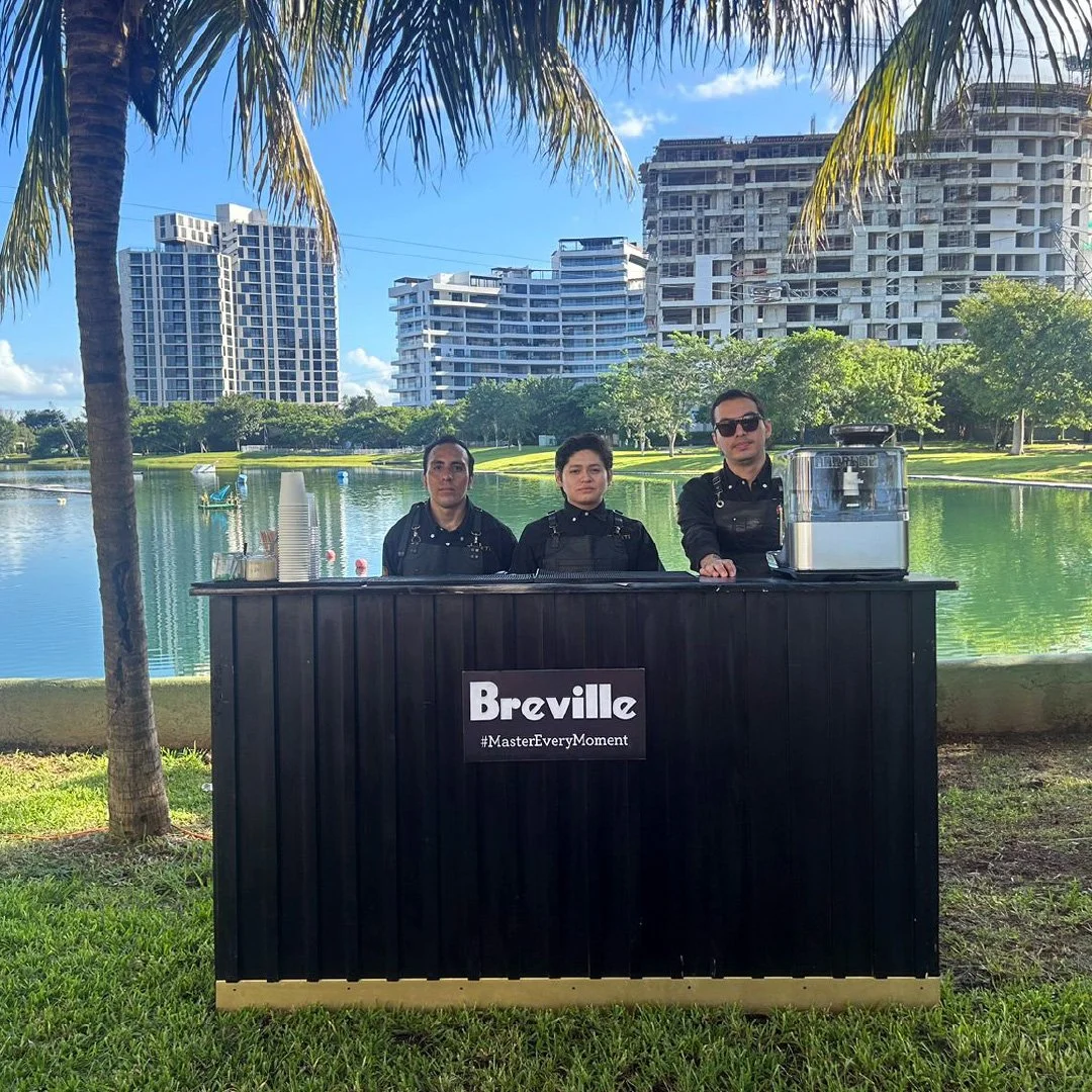 Tres personas detrás de un puesto de café Breville, con un fondo de lago y edificios residenciales, con árboles y césped en primer plano.