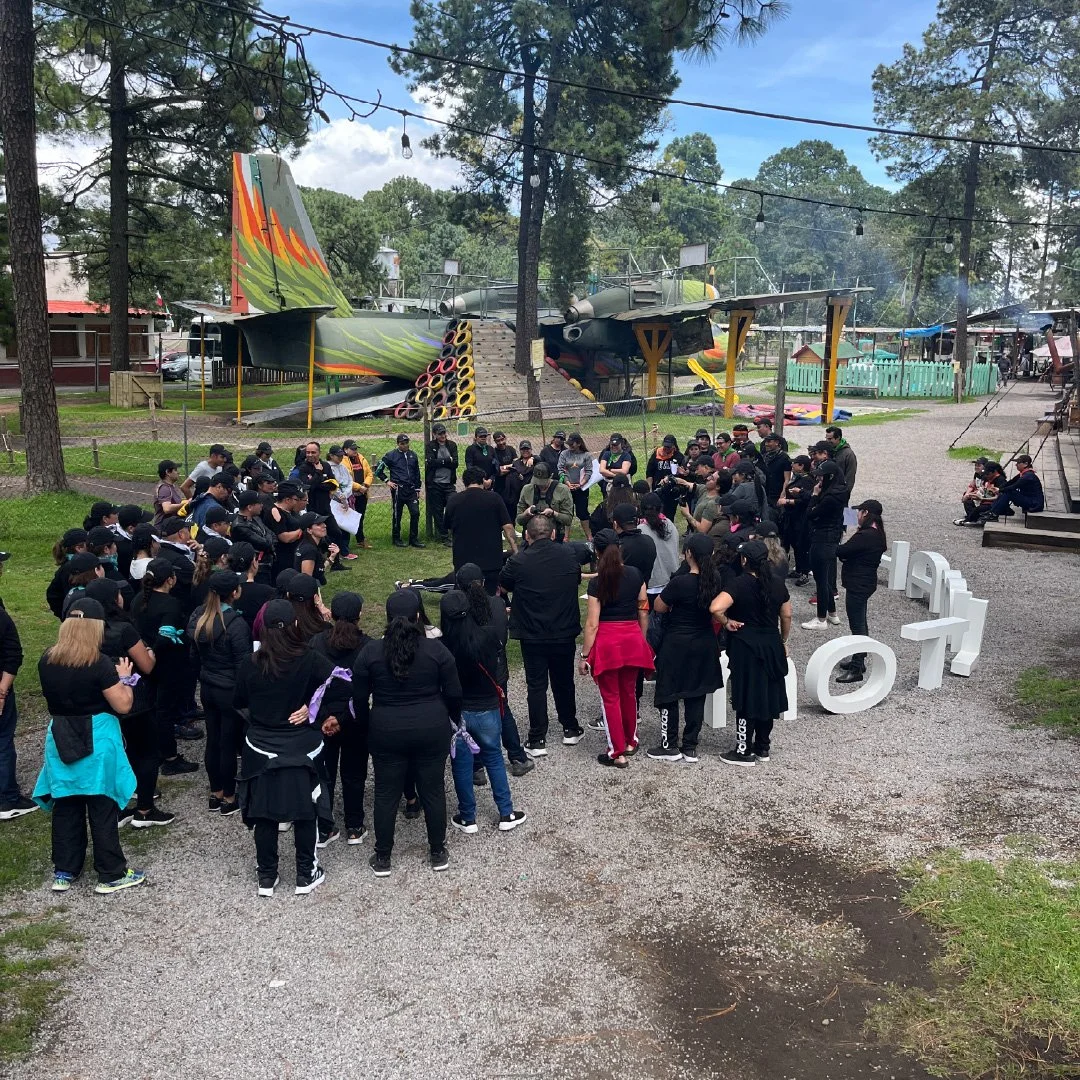 Grupo de personas reunidas frente a dos aviones de guerra en un parque, con árboles y estructuras de comida al fondo.