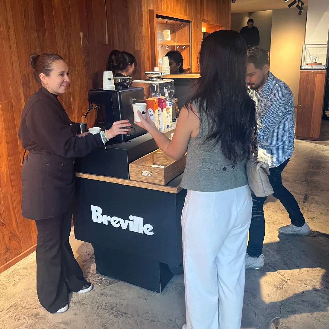 Mujer entregando un café a otra persona en un mostrador de café con la marca Breville, en un ambiente interior con paredes de madera.