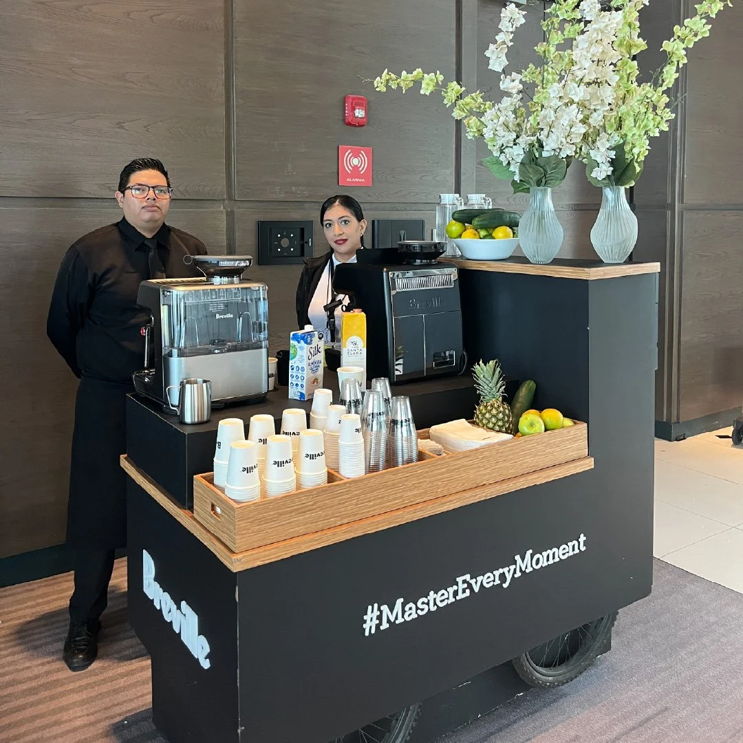Modulo de café con dos baristas en uniforme negro, con máquina de café, fruta y vasos en un entorno moderno.