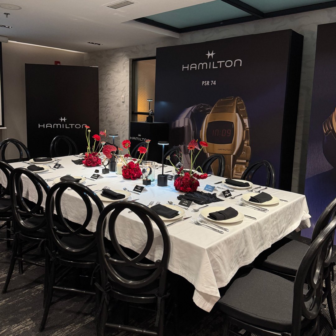 Una mesa de conferencia decorada con flores rojas y configurada con plato, cubiertos, copas y servilletas negras en un ambiente de evento, con anuncios publicitarios de Hamilton para relojes en el fondo.
