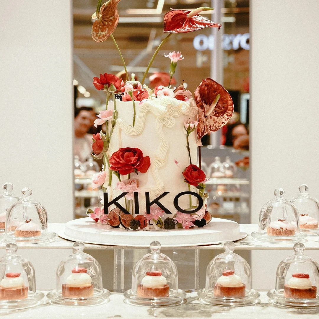 Pastel de cumpleaños decorado con flores y hojas en tonos rojos, rosas y verdes, con letras que dicen 'KIKO MILANO' en la parte frontal, rodeado de pequeñas tartas con glaseado y decoradas con flores, en un ambiente de celebración