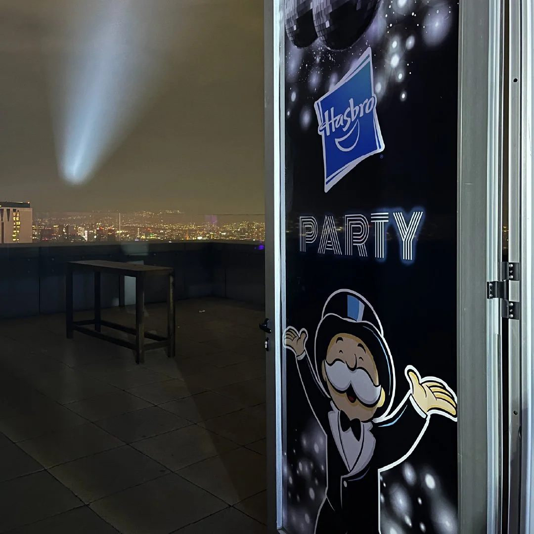 Puerta con cartel que dice 'Hasbro Party' y muestra una figura de un personaje con sombrero y bigote, en un espacio en la azotea con vista a la ciudad por la noche, con iluminación y humo en el fondo.