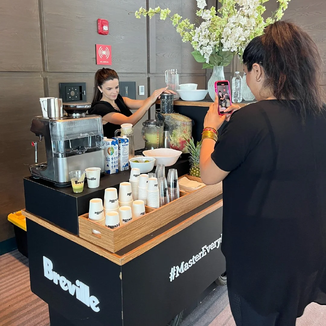 Una mujer sirviendo bebidas en un puesto de café con un cartel que dice #MasterEveryday, otra mujer tomando una foto con su teléfono móvil, y múltiples tazas, complementos y fruta en el mostrador.