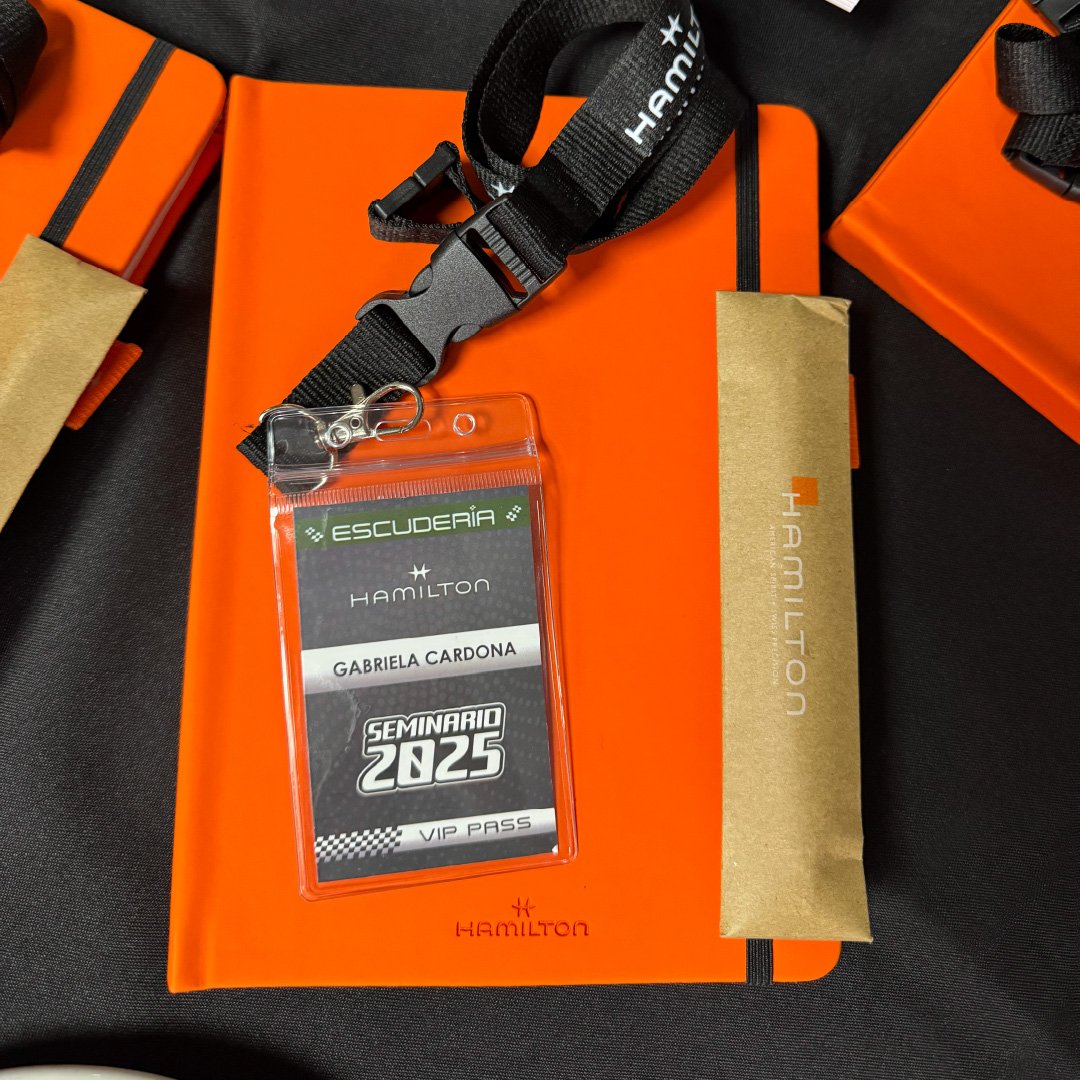 Credencial de acceso al seminario Hamilton 2025 con nombre Gabriela Cardona, en una funda de plástico transparente. Al fondo, varias cajas y accesorios de color naranja y negro, incluyendo un llavero y una caja de embalaje.
