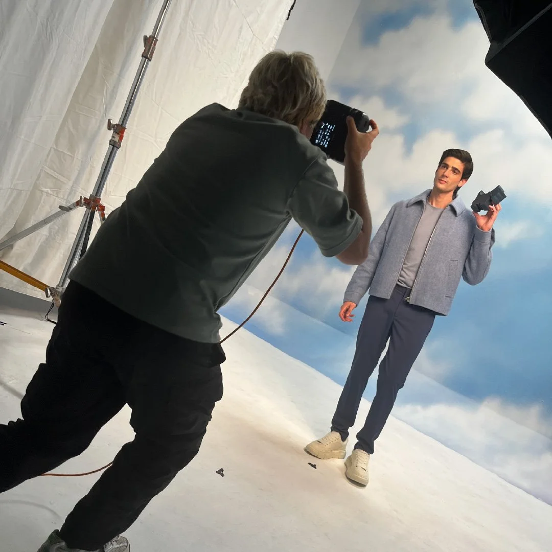 Un hombre de pie sosteniendo un teléfono mientras otra persona toma una foto en un estudio de grabación con fondo de nubes y cielo azul.