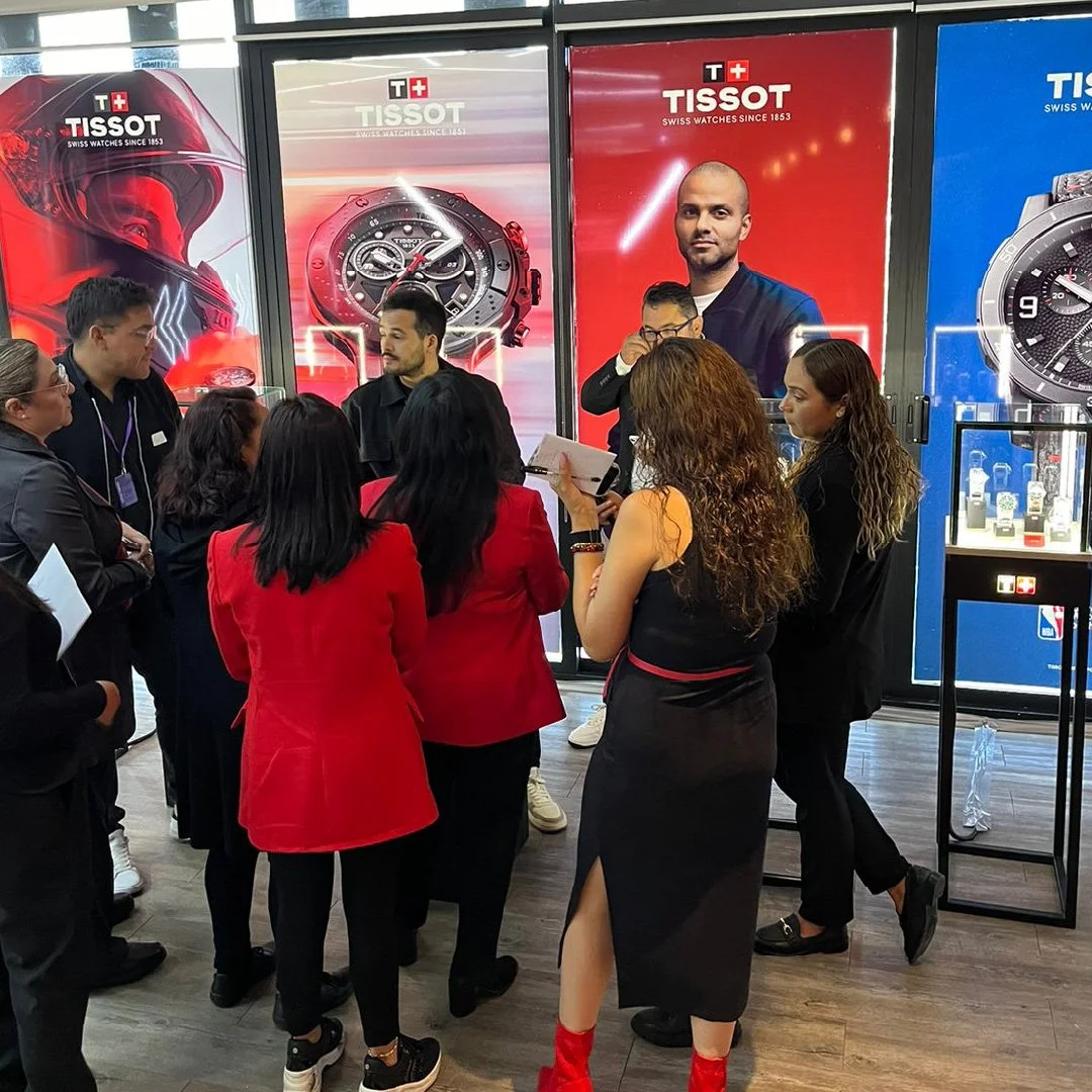 Grupo de personas en una exhibición de relojes Tissot, algunas vestidas con chaquetas rojas, otras en ropa negra, conversando y tomando notas, con cartelera de relojes en el fondo.