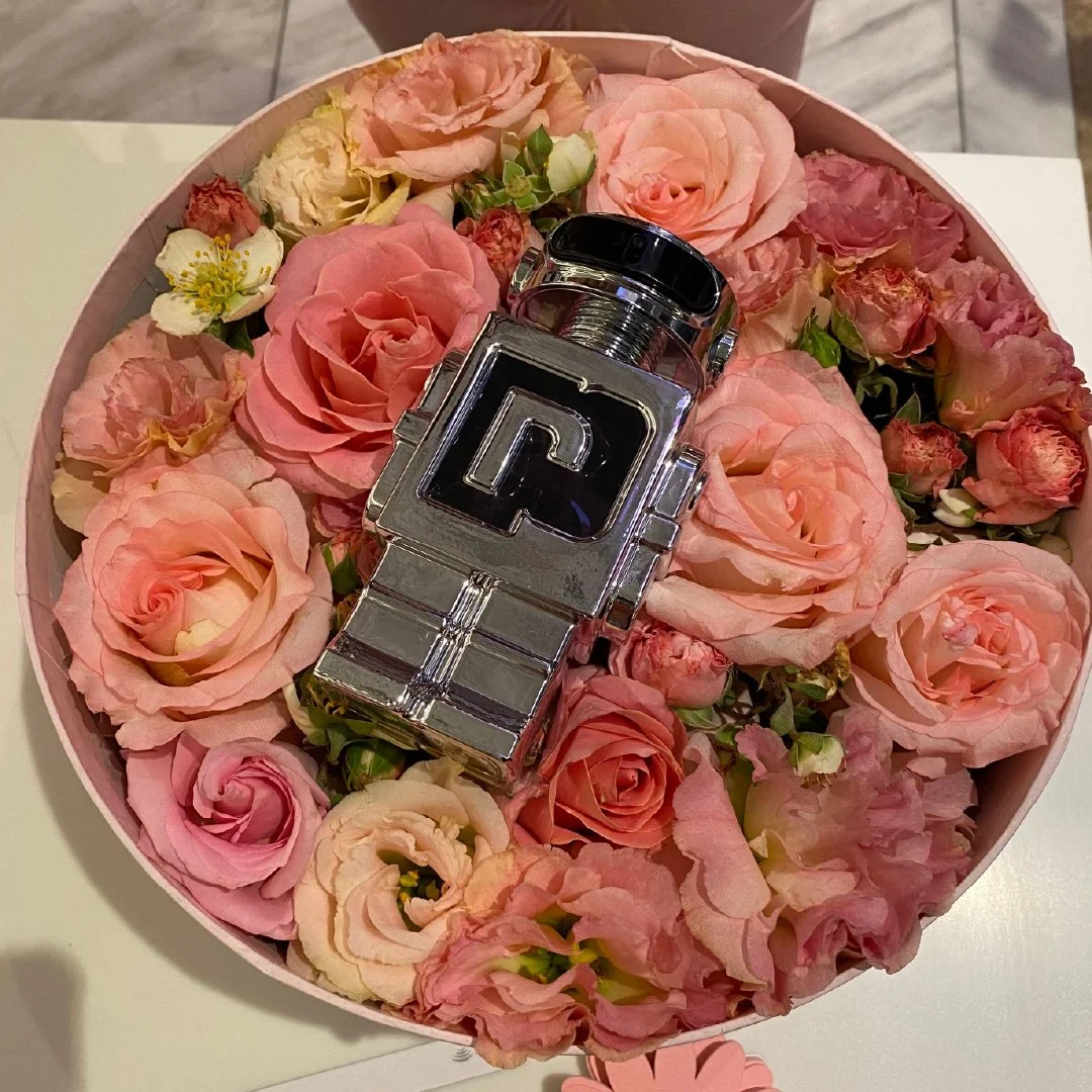 Un frasco de perfume plateado sobre un arreglo de flores rosas y durazno en una caja redonda