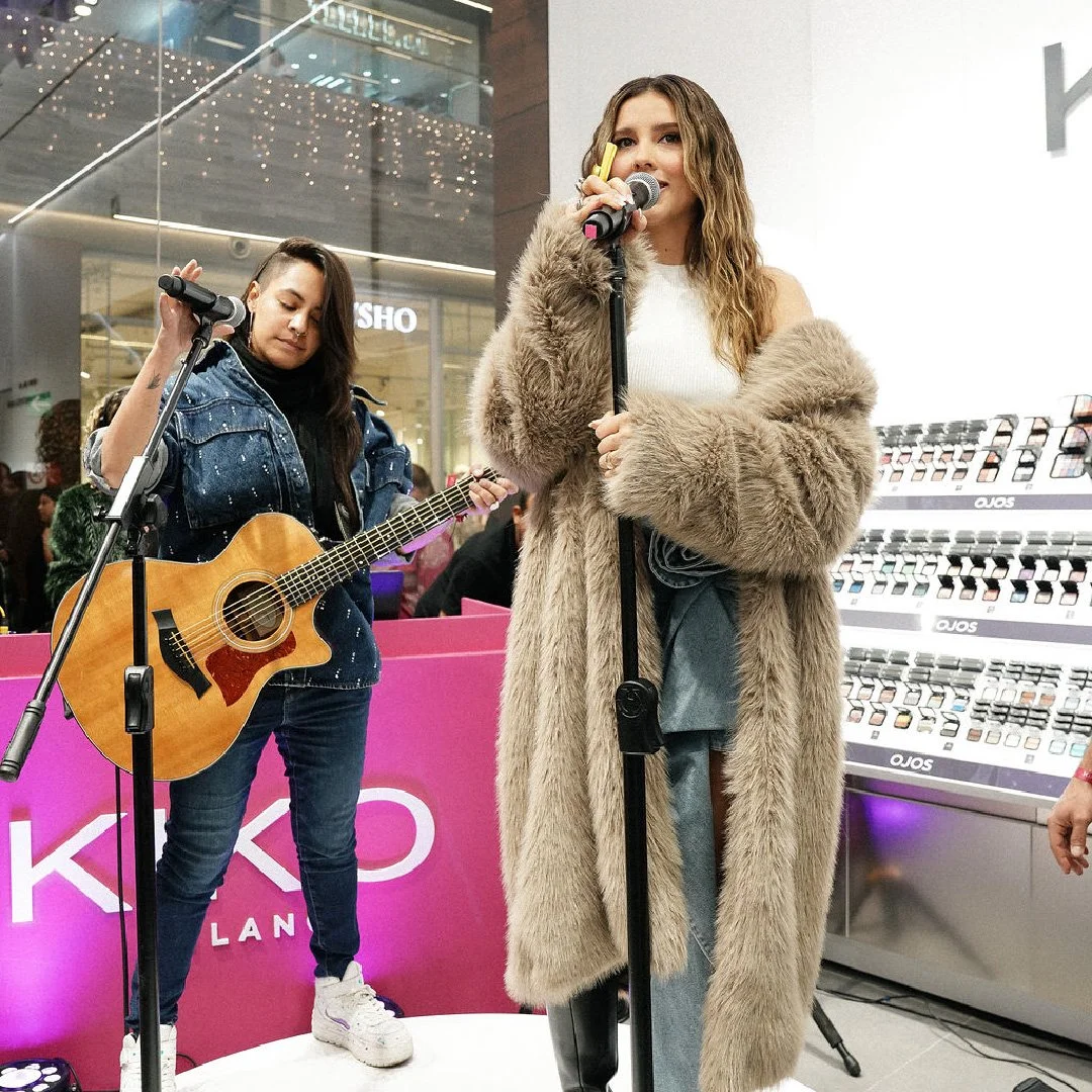 Una mujer cantando con un micrófono, lleva un abrigo de piel y un conjunto gris, junto a ella hay otra mujer tocando una guitarra en un escenario en un centro comercial.