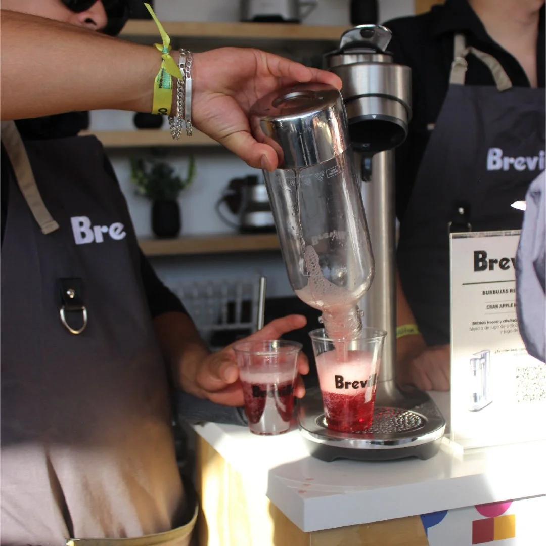 Persona sirviendo una bebida roja y espumosa en un vaso transparente con la marca "Brevi" en una máquina de bebidas en un entorno de cafetería.