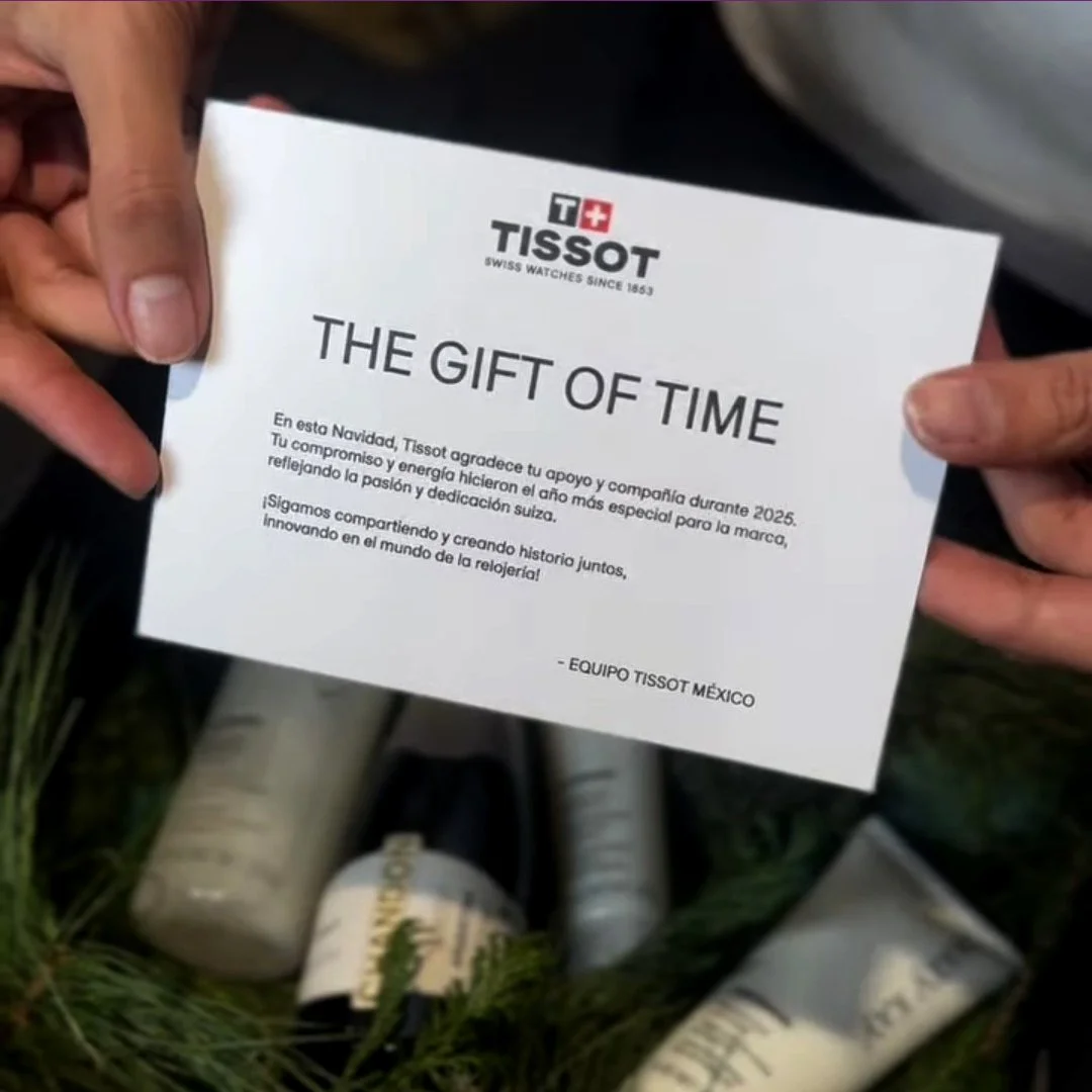 Mano sosteniendo una carta con el logo de Tissot, mensaje de agradecimiento por apoyo en 2025, en fondo con productos de la marca en medio de vegetación.