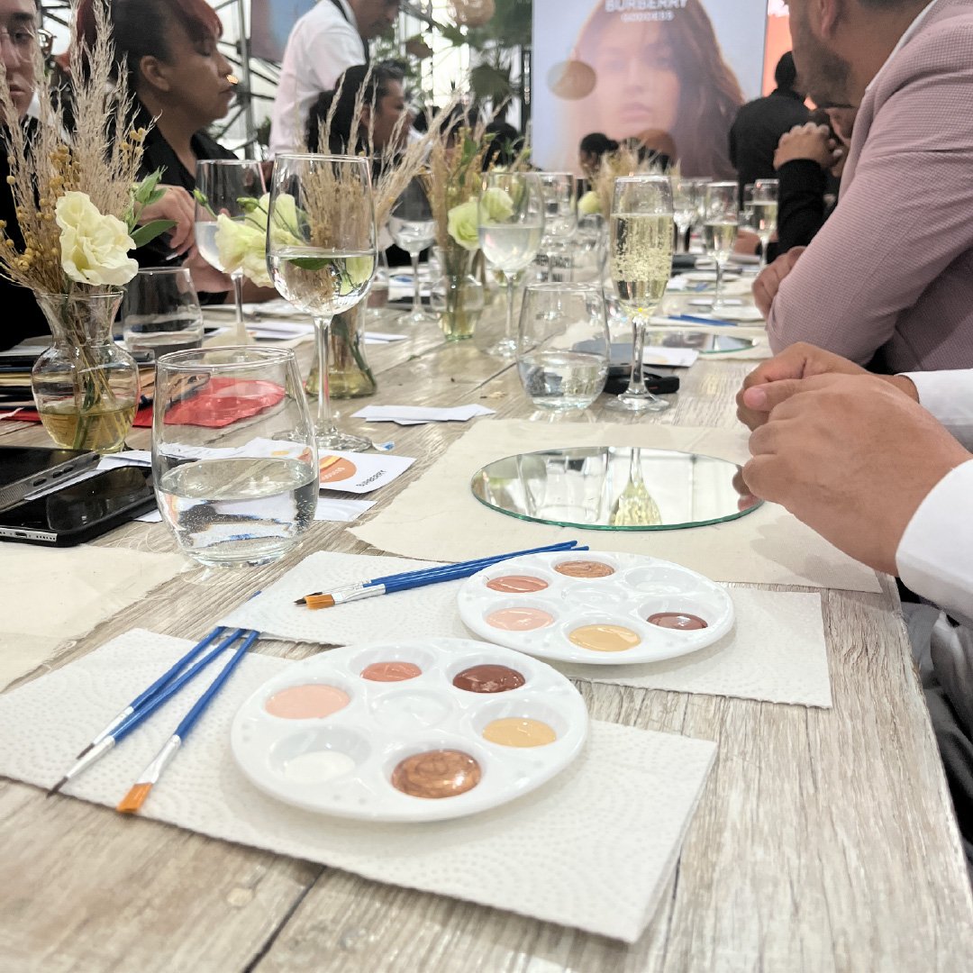 Mesa de reunión con galería de acuarelas y pinceles, varias copas de agua y vino, y decoraciones florales.