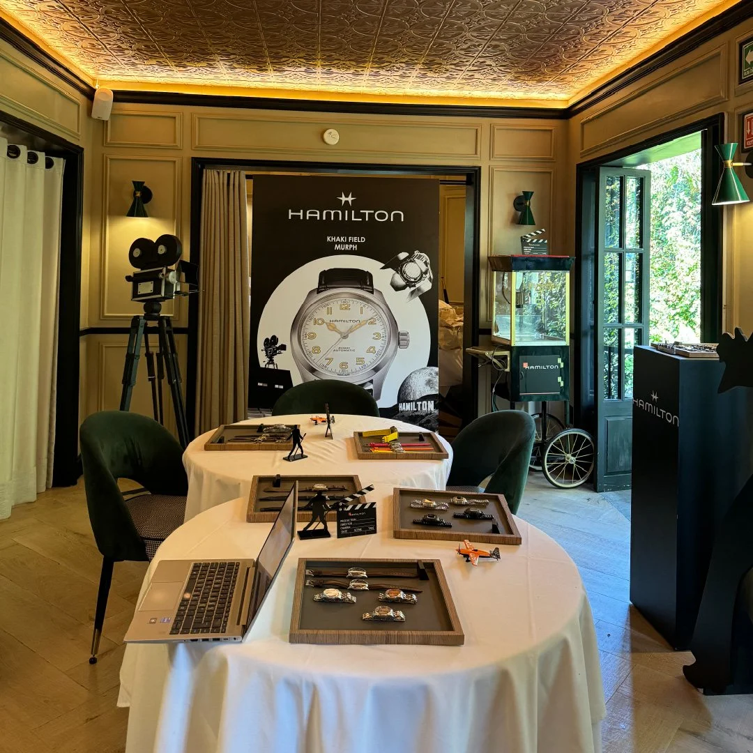 Muestra de relojes en una mesa, con una cabina de videograbación y un cartel grande de Hamilton en el fondo.