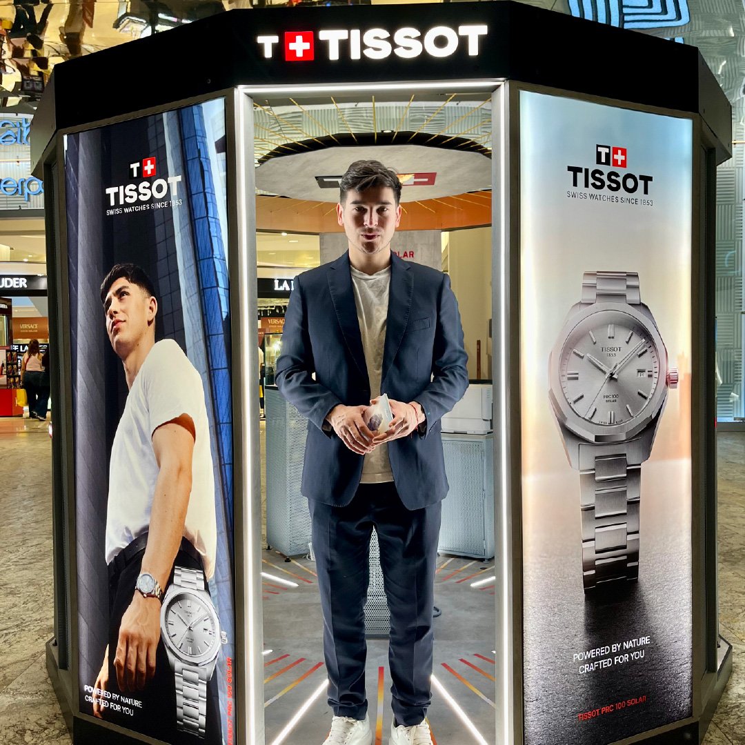 Hombre joven vestido con traje oscuro, de pie en una cabina de publicidad para relojes Tissot en un centro comercial, con anuncios de relojes en los lados.