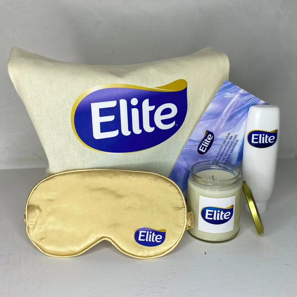 Set de productos de higiene con la marca Elite, que incluye una almohada de tela, una máscara para dormir de satín dorado, una vela en frasco, un gel o crema en envase rectangular y un spray, todos con etiquetas de la marca Elite.