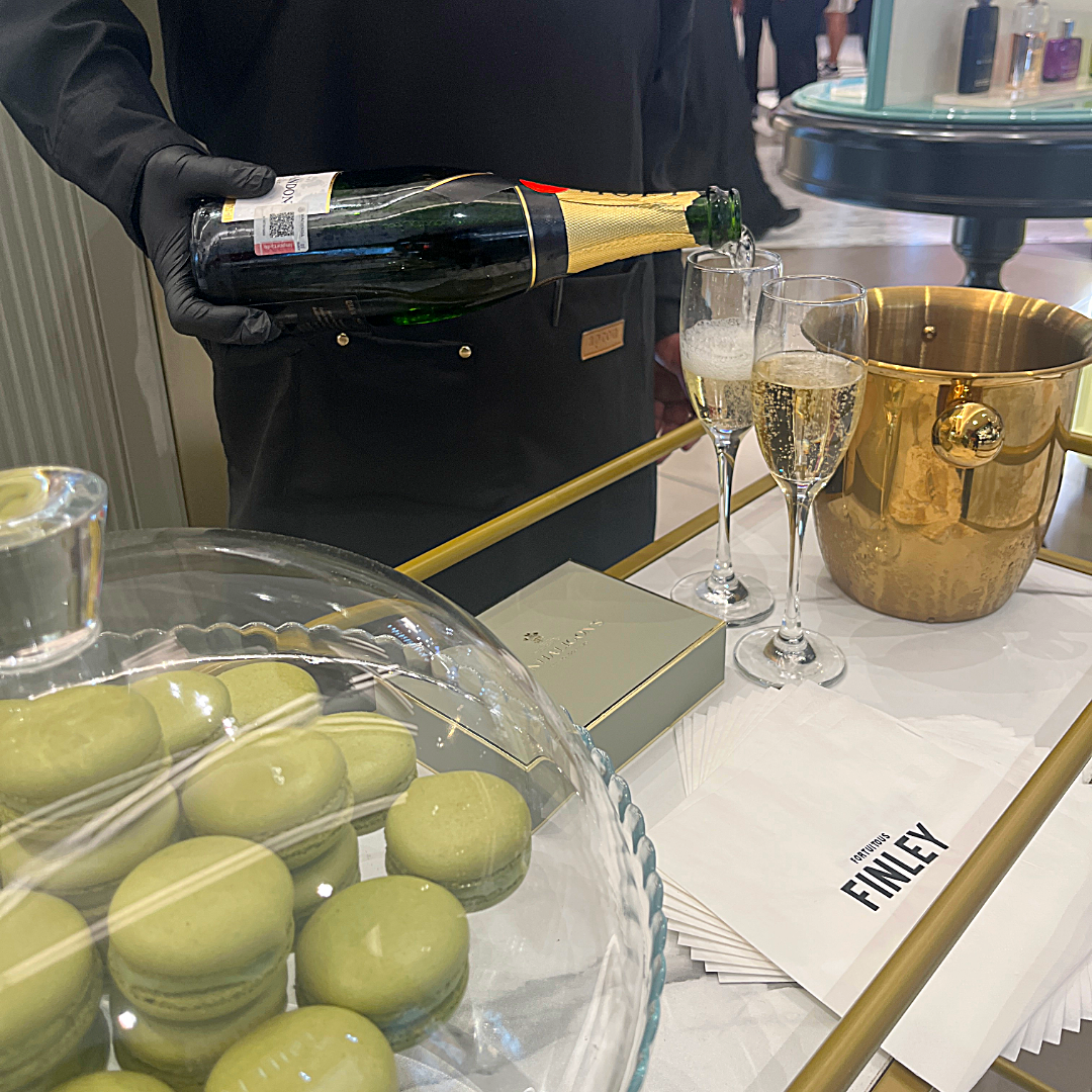 Persona sirviendo champán en copas junto a una bandeja de macarons verdes y una caja de finley en una mesa decorada con artículos de lujo.