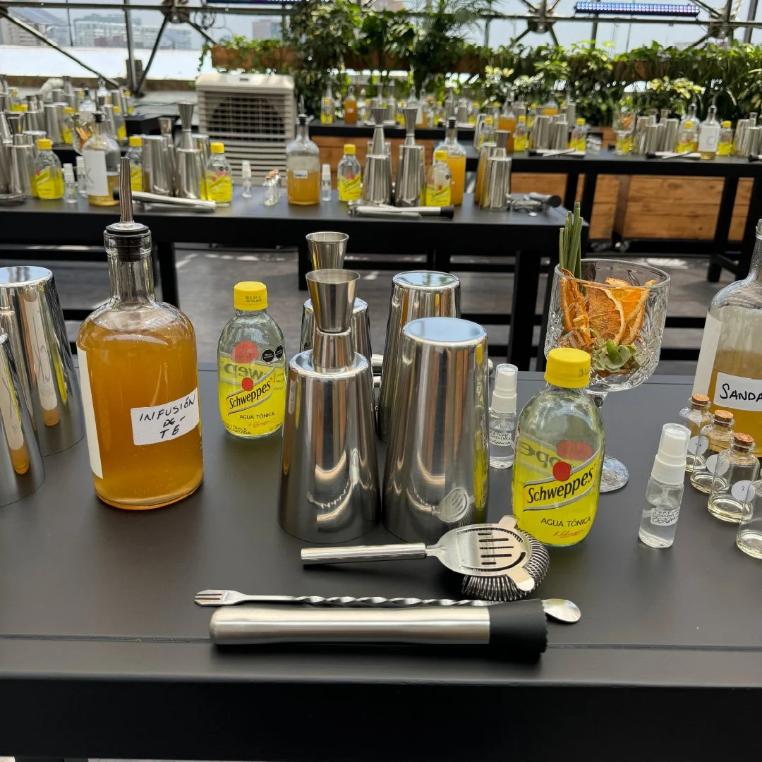 Mesa de bar con botellas de agua tónica, jarra de infusión de té, copas, mezcladores metálicos, un agitador y decoraciones con frutas secas y vegetales.