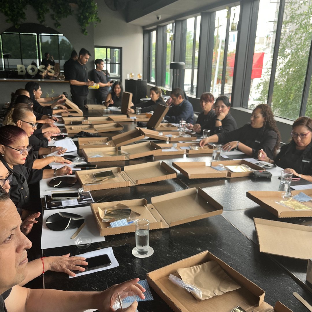 Grupo de personas en una reunión en una sala con grandes ventanales, con cajas de comida sobre la mesa y algunos asistentes usando teléfonos móviles.