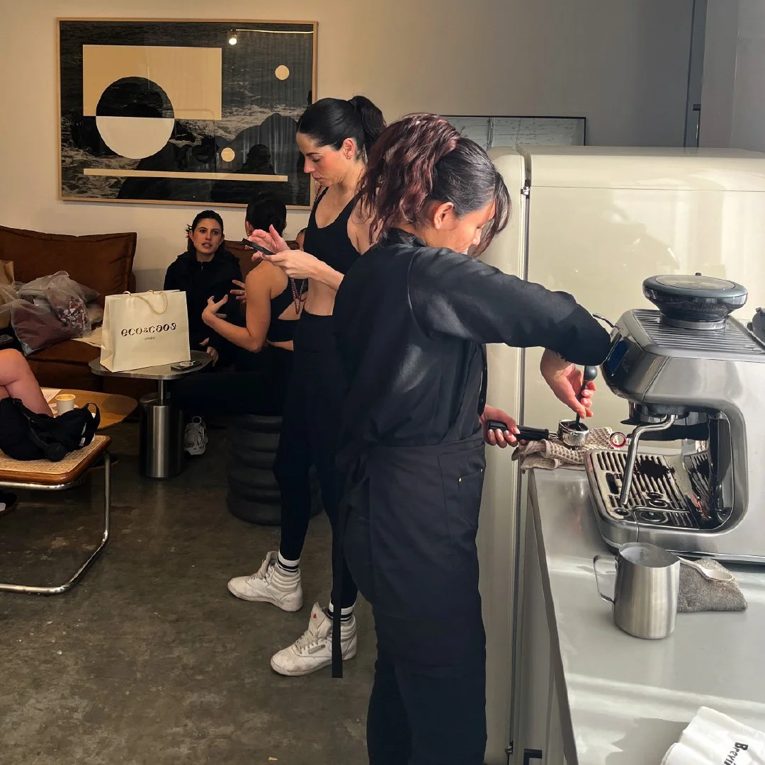 Dos mujeres preparando café en una cafetería y otras tres sentadas en un sofá conversando.