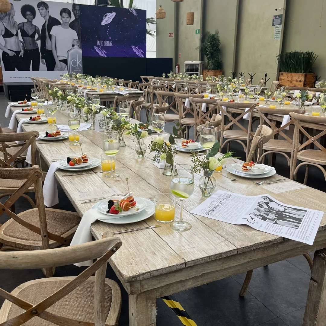 Mesa decorada para evento con platos, vasos con jugo y vino, flores y anuncios en un espacio abierto.