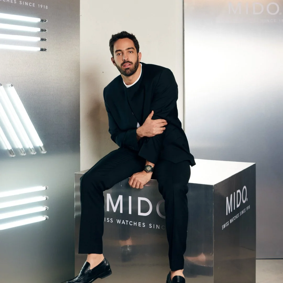 Un hombre con barba viste un traje negro y está sentado en una caja con la marca MIDO, en una tienda o exhibición de relojes.