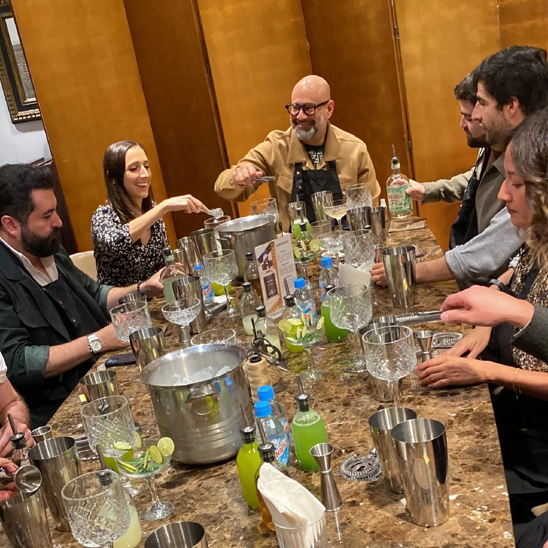 Grupo de personas en una mesa de bar cocktail, preparando y sirviendo bebidas alcohólicas, con un bartender en el centro.
