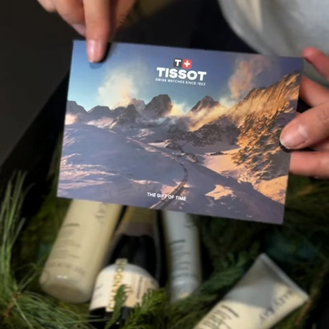 Mano sosteniendo un folleto de la marca Tissot con una imagen de montañas nevadas y un camino en medio de ellas.