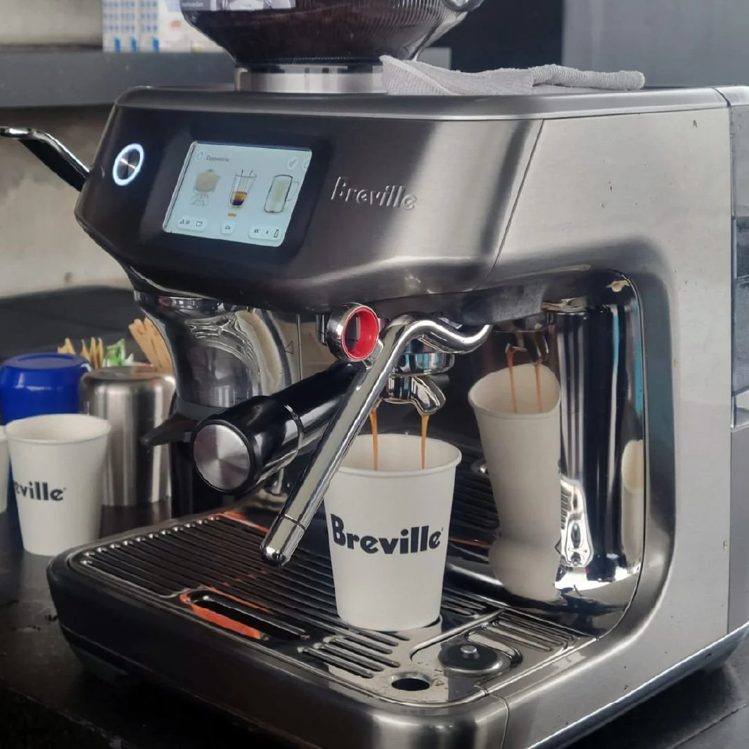 Máquina de café espresso vertiendo café en varias tazas de cartón con el logo 'Breville'.