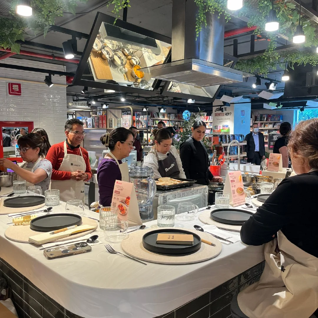 Grupo de personas en un taller de cocina en un espacio moderno y bien iluminado, con estantes llenos de libros y plantas decorativas en el fondo.