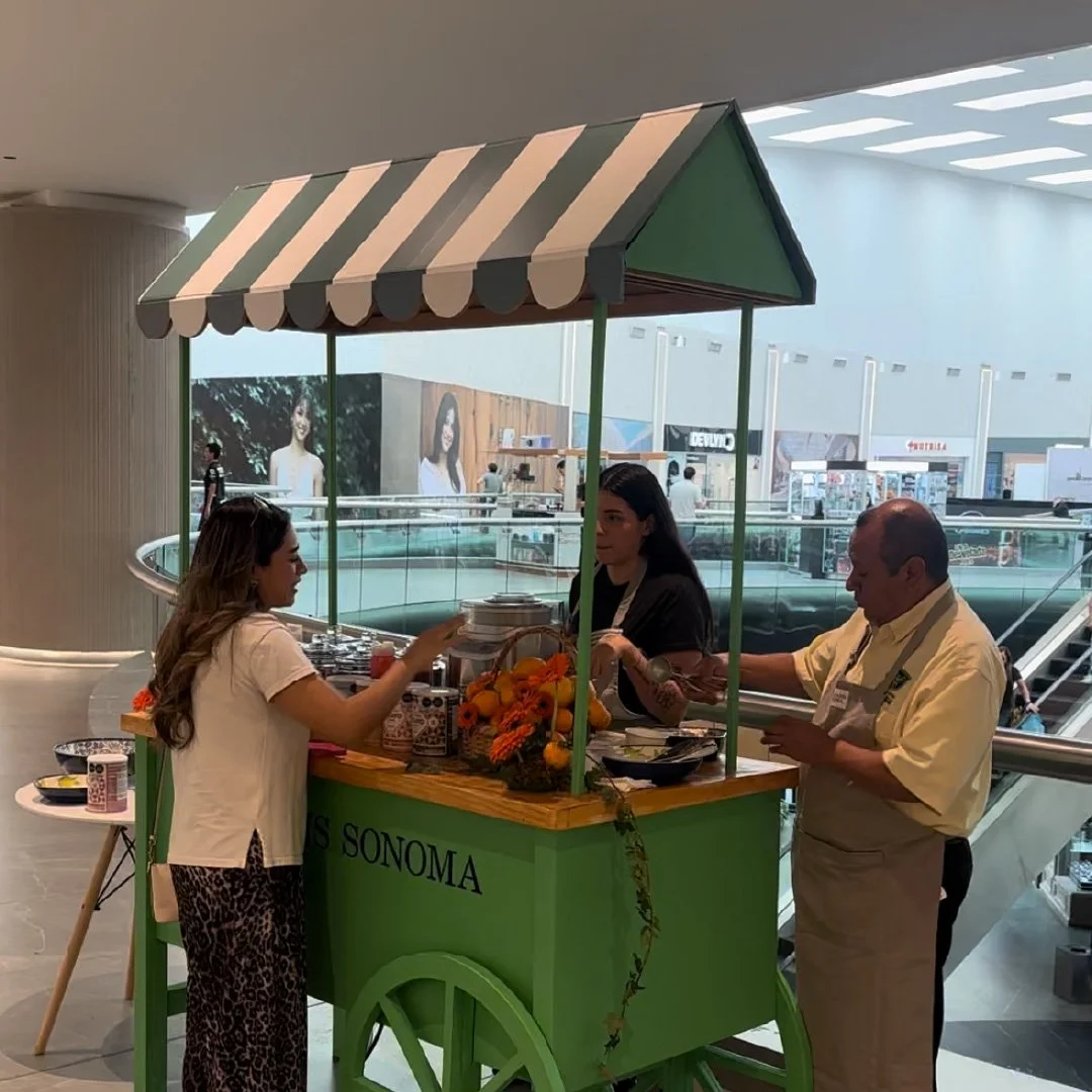 Carrito de nieve en un centro comercial, con dos personas atendiendo a una clienta. El carrito tiene un toldo a rayas verde y blanco, y está decorado con flores naranjas y verdes. williams sonoma
