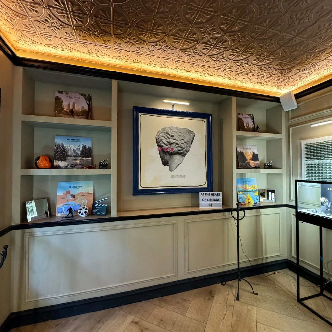 Una sala de exhibición con estanterías blancas que contienen libros y objetos decorativos, un marco con una escultura de la cabeza de una figura clásica adornada con flores, y una pantalla en un soporte negro en la esquina derecha de la imagen.