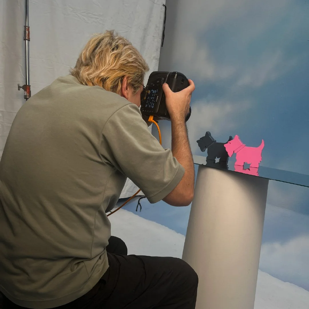 Persona tomando una fotografía de dos figuritas de perros de papel en un fondo azul con nubes.