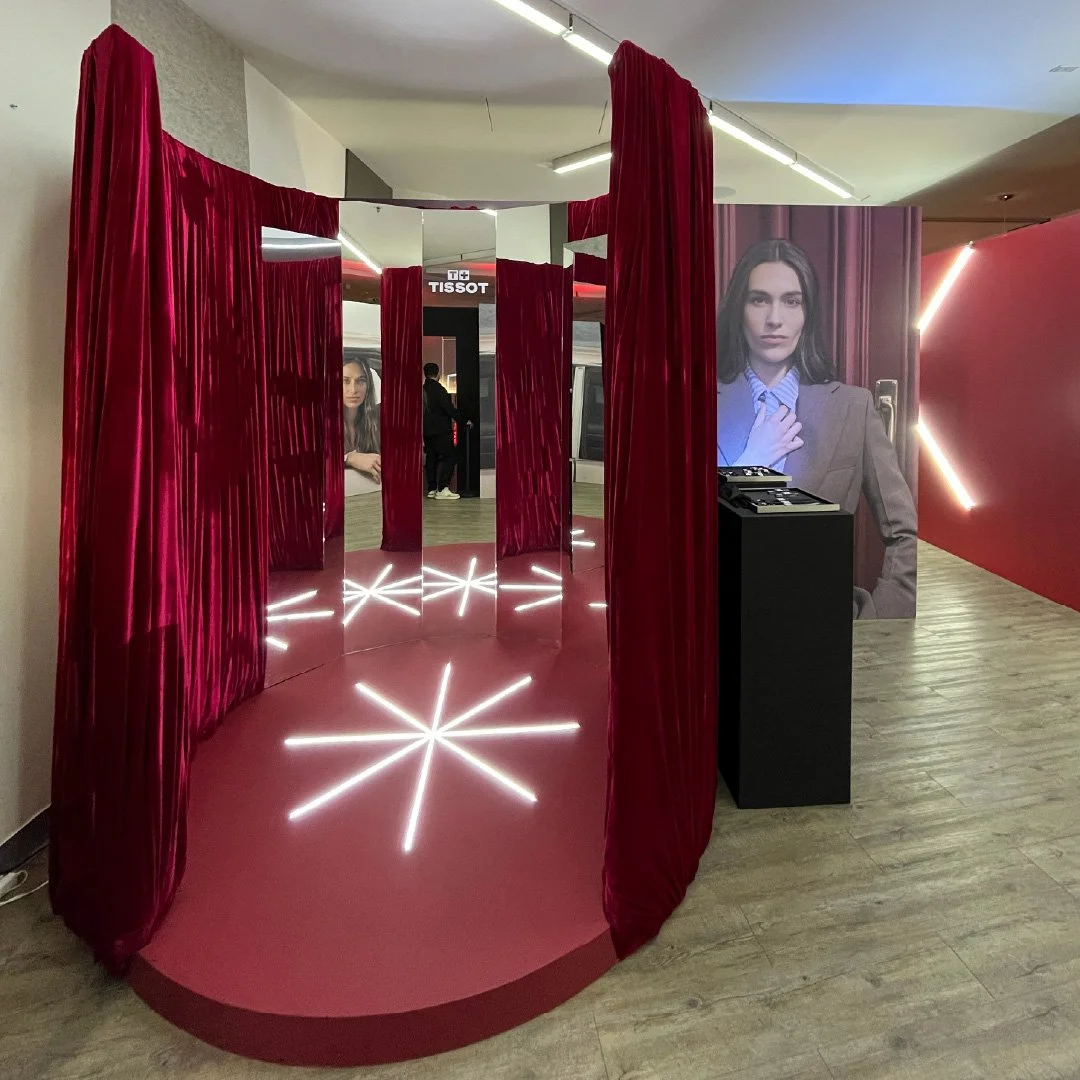 Una estructura circular con cortinas rojas y un suelo iluminado con líneas blancas que forman estrellas. En el fondo hay una pared con un retrato grande de una mujer y un stand con la marca Tissot.
