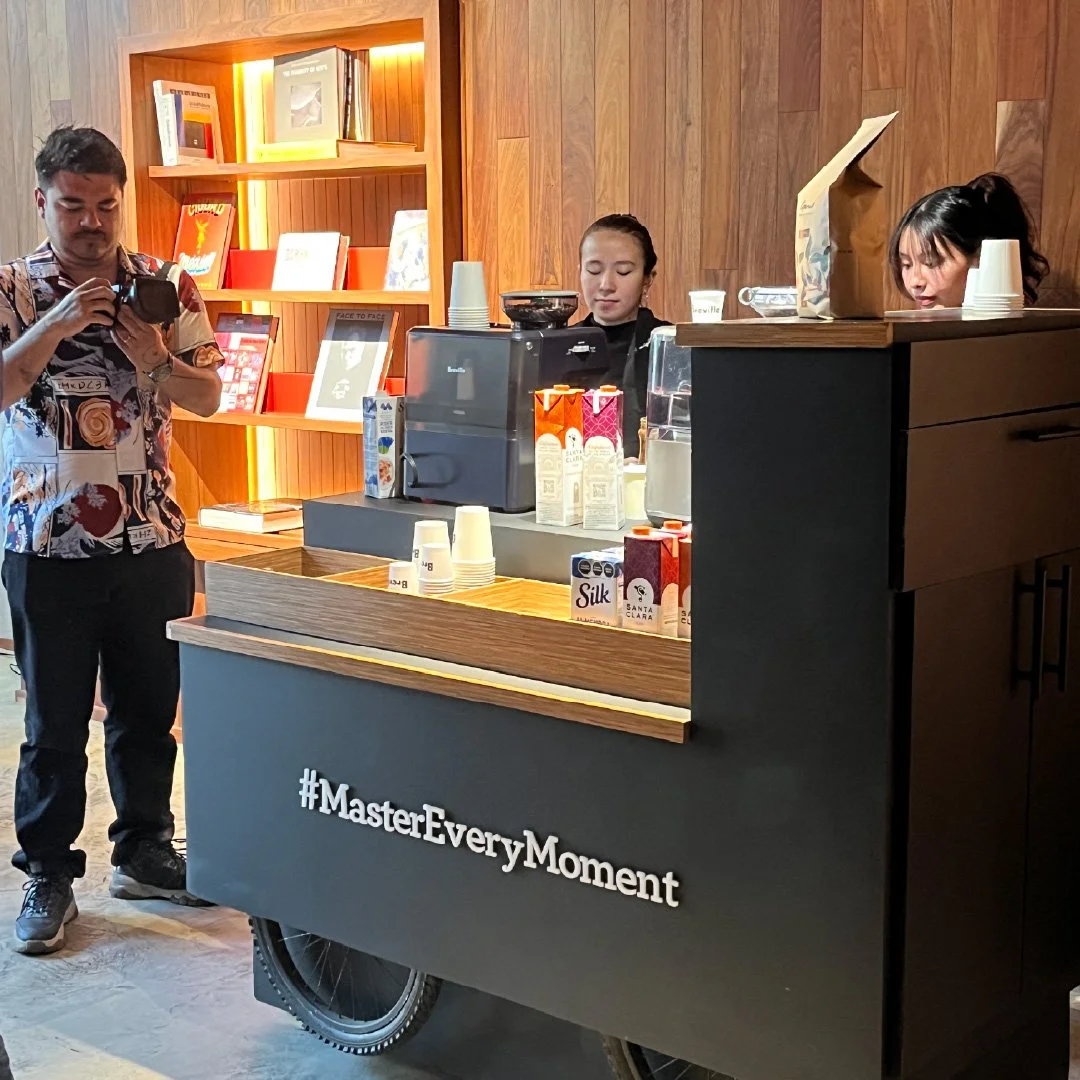 Un carrito de cafetería con dos baristas atendiendo, y un cliente tomándose una foto. La barra tiene bebidas y paquetes de leche, y en la parte frontal se lee el hashtag #MasterEveryMoment. En el fondo hay libreros de madera con libros y decoraciones
