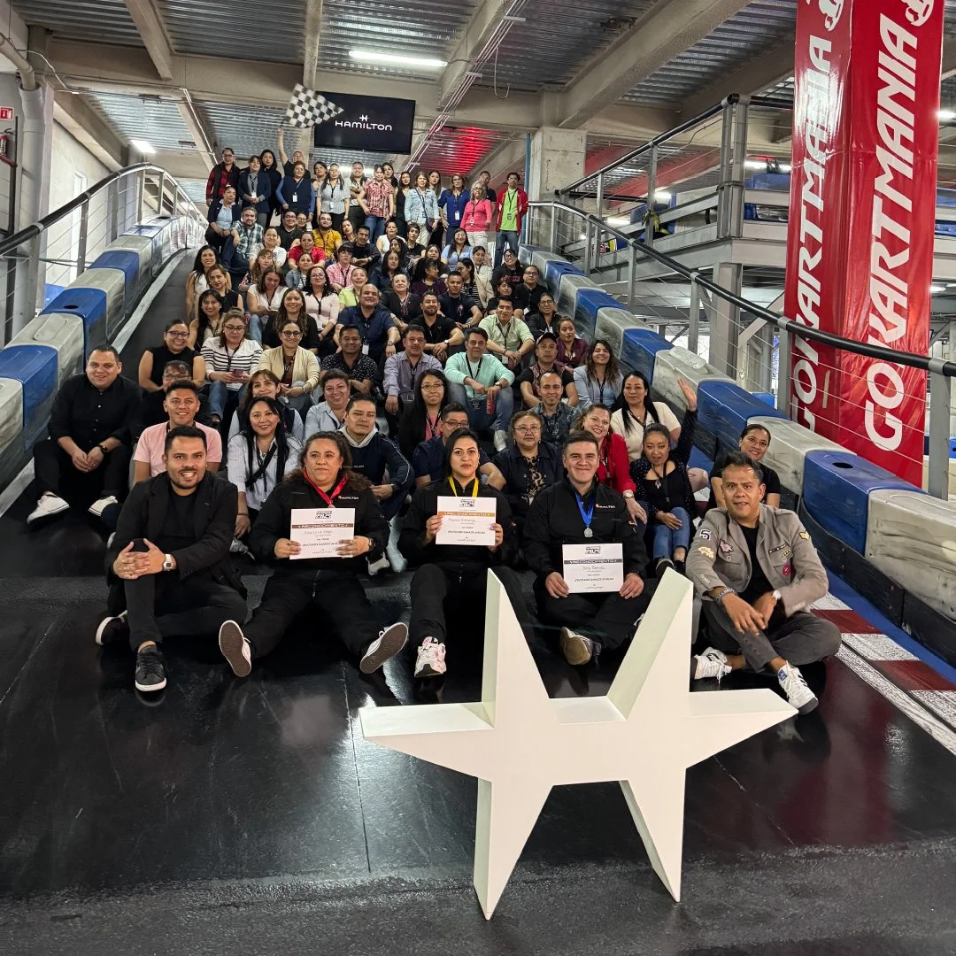 Un grupo grande de personas en una especie de estadio o arena, algunos sosteniendo certificados, en un evento con banderas de la Fórmula 1 y una estrella grande de color blanco en el frente.