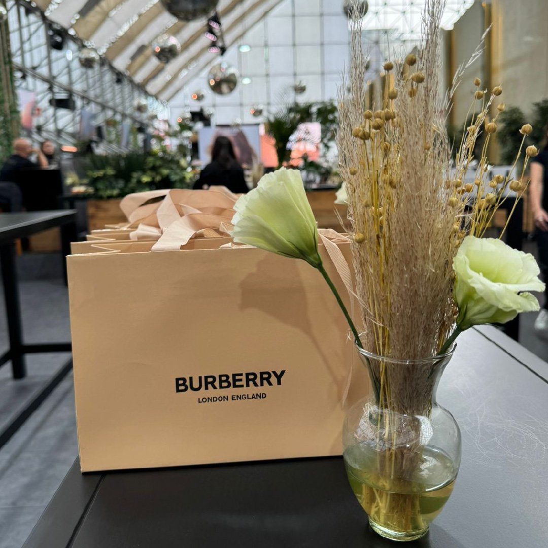 Una caja beige con la palabra 'BURBERRY' y 'LONDON ENGLAND' en letras negras, junto a un jarrón con flores secas y blancas en un ambiente de interior con personas en el fondo.