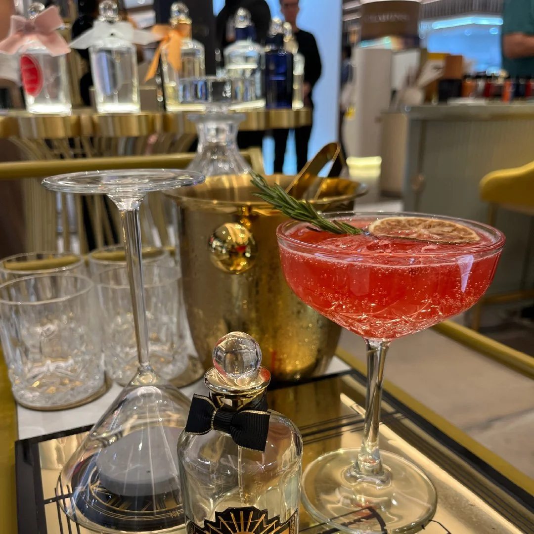 Copa de cóctel rosado con rodaja de limón y ramita de romero encima y varias botellas de perfume, vasos, y utensilios en un bar o restaurante elegante.