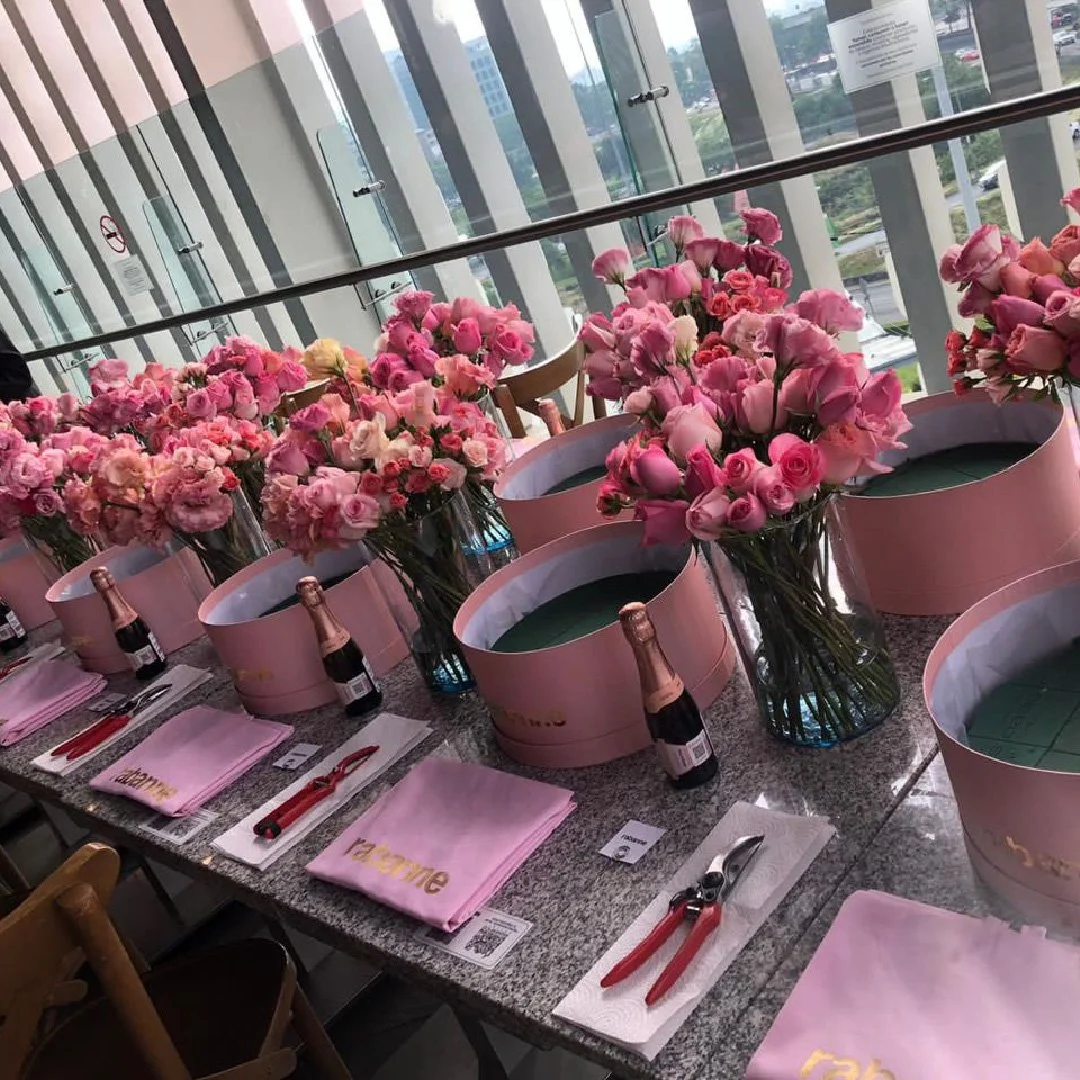 Mesas decoradas con grandes jarrones de flores rosas y botellas de champagne, con manteles rosados y utensilios listos en un espacio con ventanas grandes.