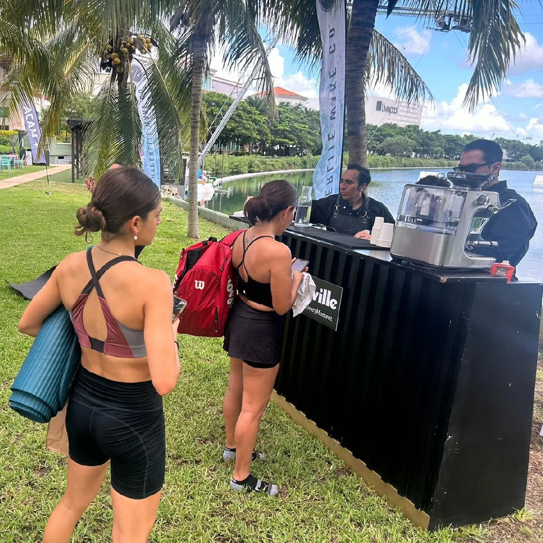 Dos mujeres hacen fila en un stand de café al aire libre cerca de un lago, con palmeras y árboles en el fondo.