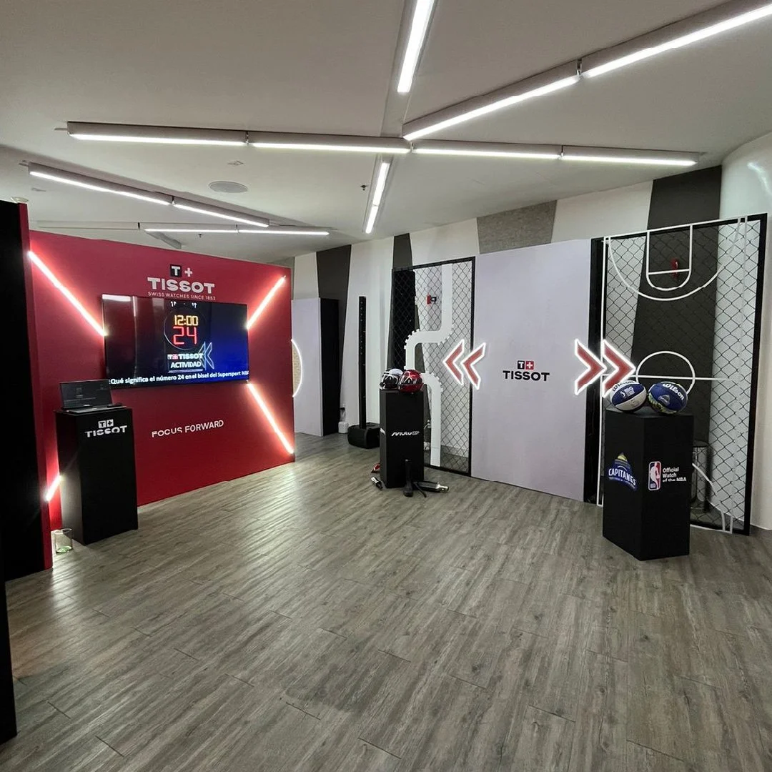 Espacio interior con decoración deportiva y de la marca Tissot, con una pantalla digital, balones de baloncesto y cascos en exhibición.