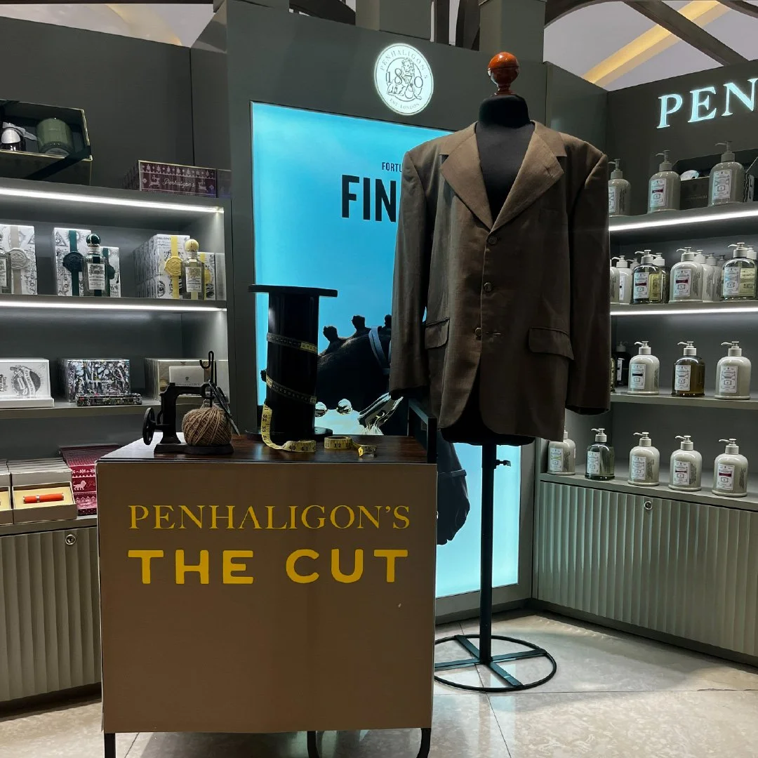 Un puesto de exhibición que muestra un maniquí vestido con un saco marrón, junto a un cartel que dice 'Penhaligon's The Cut'. En la mesa hay un carrete de hilo, una cinta métrica, y una máquina de coser miniatura. En las estanterías se ven botellas d