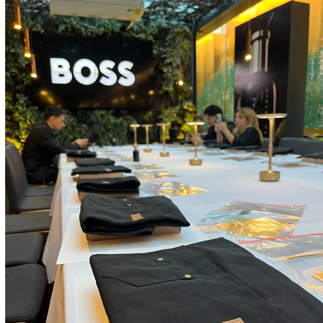 Mesa larga en un restaurante con prendas negras bien dobladas y bolsas plásticas con prendas. En el fondo, paredes verdes con plantas y un letrero luminoso que dice 'BOSS'.