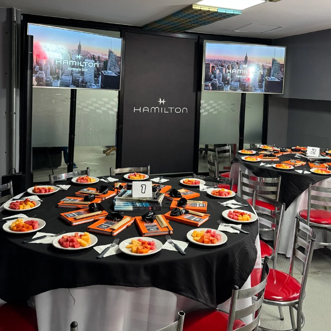 Sala de reuniones con mesas redondas cubiertas con manteles negros y platos con fruta picada. En el centro de la mesa hay folletos, audífonos y un cartel con número 1. En la pared hay dos pantallas que muestran una ciudad y el texto 'Hamilton Seminar