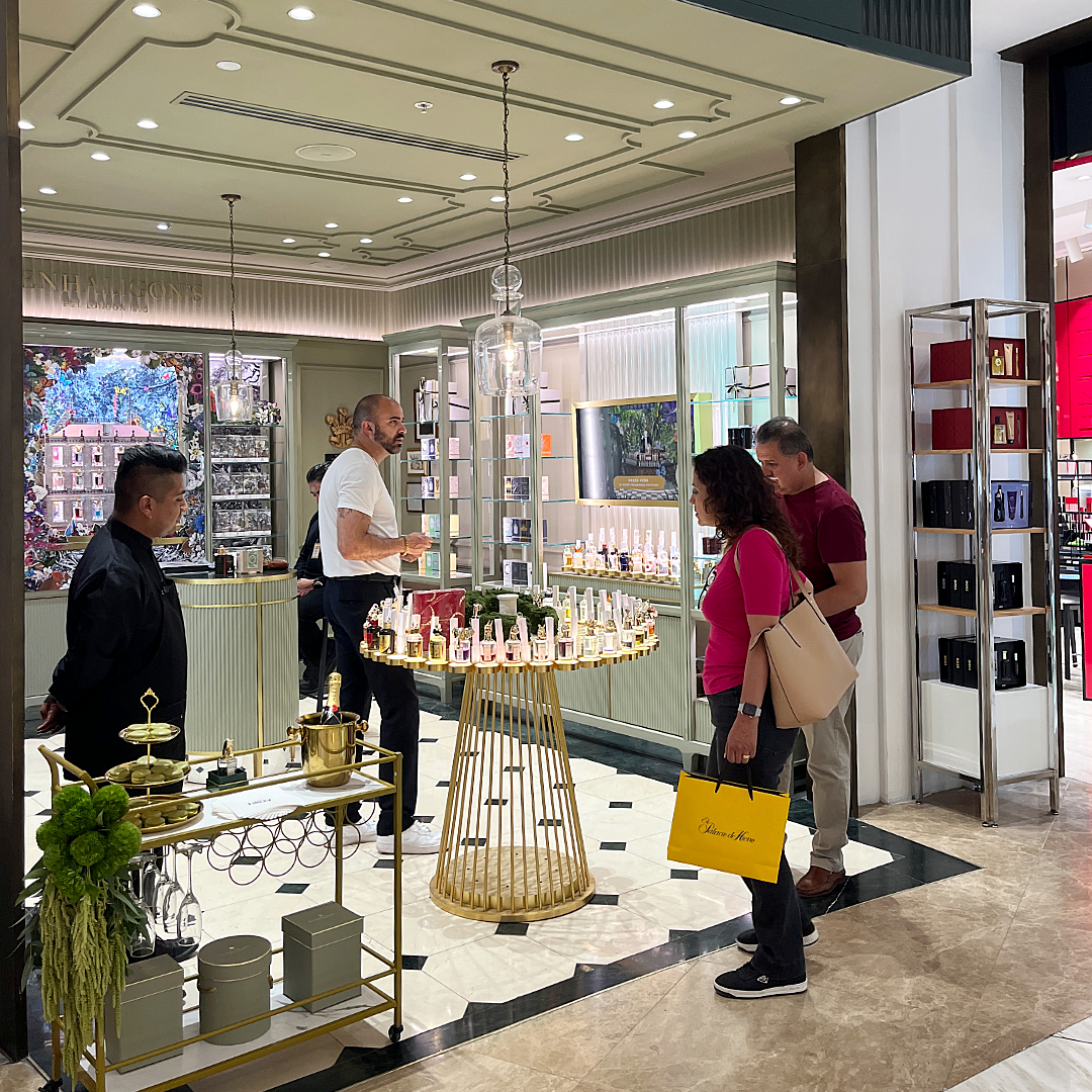 Clientes en una tienda de cosméticos y fragancias, con un empleado atendiendo en el mostrador y estantes con productos en exhibición.