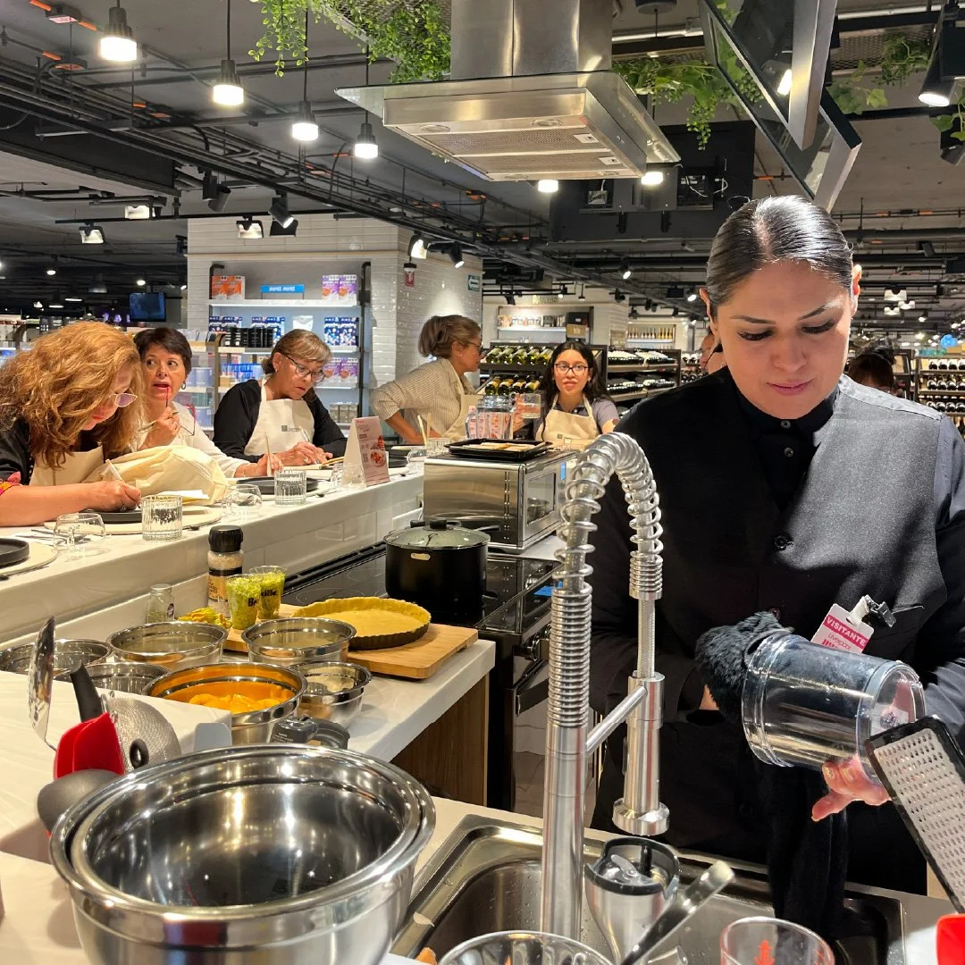 Grupo de mujeres en un curso de cocina en una tienda de supermercados, con ingredientes y utensilios en la mesa, y una instructor observando atentamente.