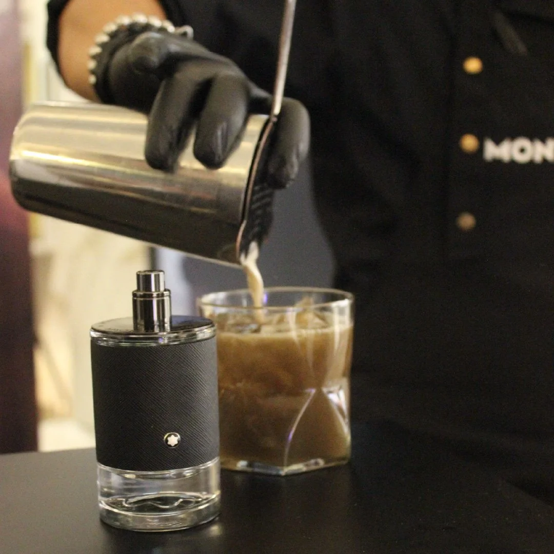 Persona sirviendo café con leche en un vaso de vidrio en una mesa, usando guantes negros y un recipiente de metal.