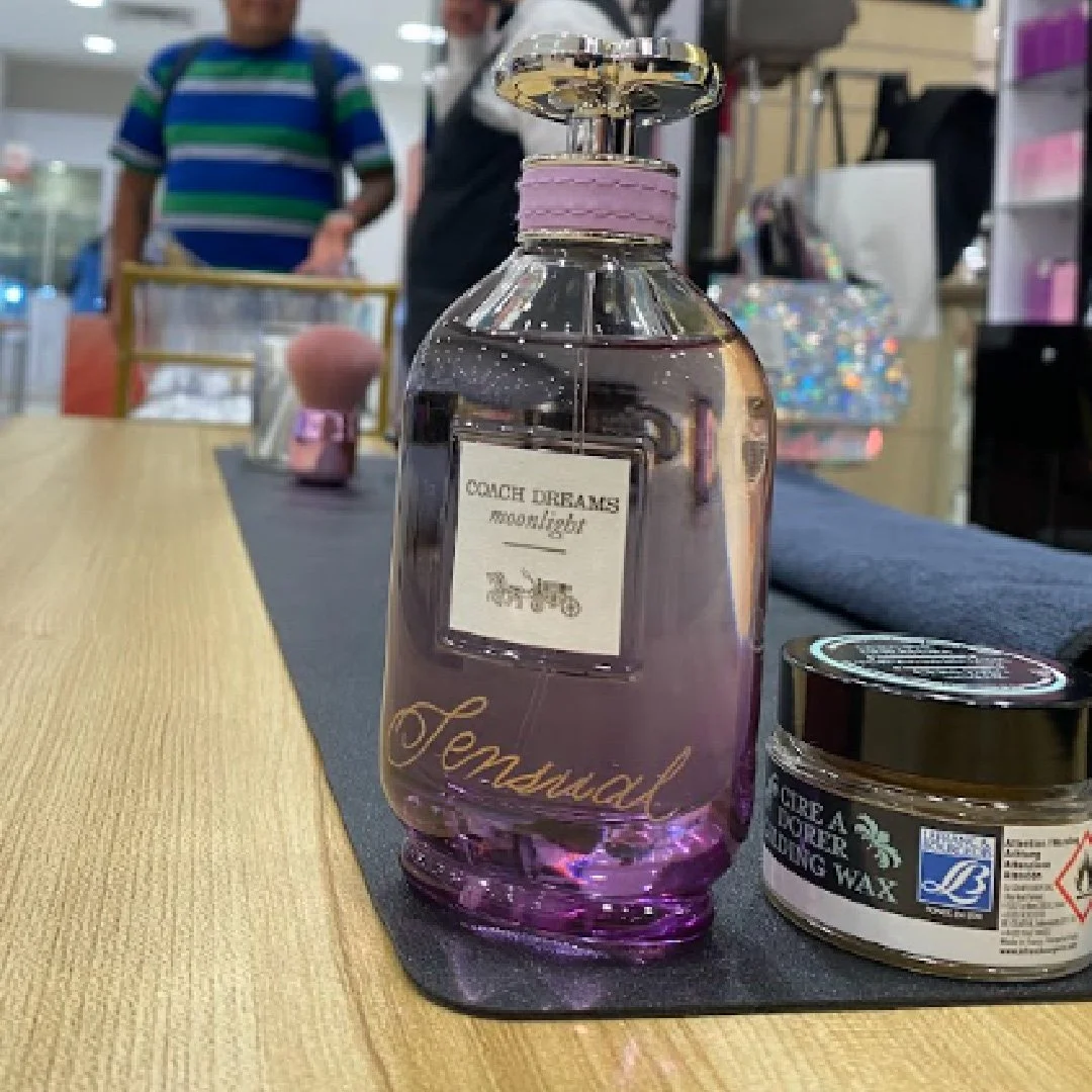 Botella de perfume de color lilas en una mesa de madera, con un frasco de cera y otros objetos en el fondo.