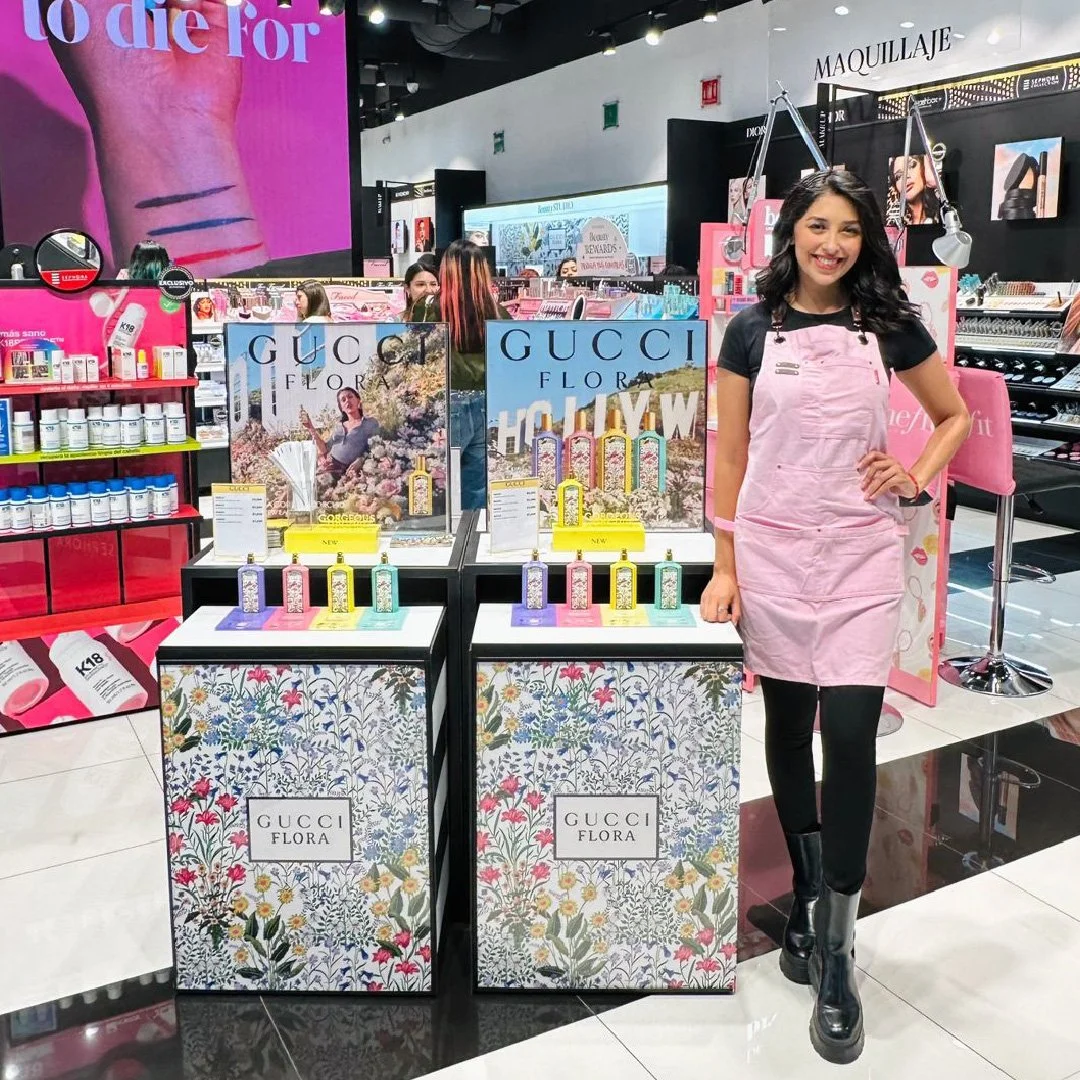 Mujer sonriendo con delantal rosa en tienda de belleza, exhibiendo productos de Gucci Flora en display floral y publicidad de maquillaje y cuidado de pieles.